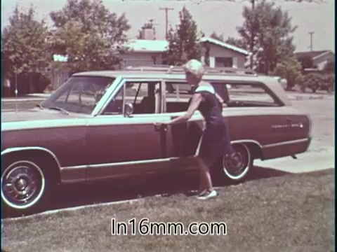 The still shows Shirley Shahan, a professional drag racer, stepping out of a vintage station wagon. She has a confident demeanor, emphasizing safety in driving. The background features suburban houses and greenery, reflecting a casual setting. This image conveys her belief in safety while engaging in motorsport, contrasting traditional views of drag racing as a rough sport for women.