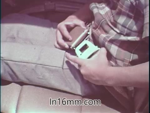 The still shows a person sitting in a vehicle, preparing to fasten a seatbelt. Their hand is gripping the seatbelt latch, while the other rests on their knee. The seat appears to be part of a car interior, emphasizing safety in automotive contexts. This moment aligns with Shirley Shahan's emphasis on safety during drag racing, highlighting a proactive attitude towards using safety measures.