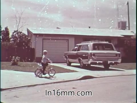 The still shows a suburban scene featuring a young boy riding a bicycle on the sidewalk, with a vintage station wagon parked in the driveway of a house. The setting suggests a typical 1960s neighborhood, reflecting a focus on family and safety, complementing the context of Shirley Shahan's message about safety in drag racing. The scene underscores the importance of safe driving, especially in residential areas, aligning with the PSA's theme.