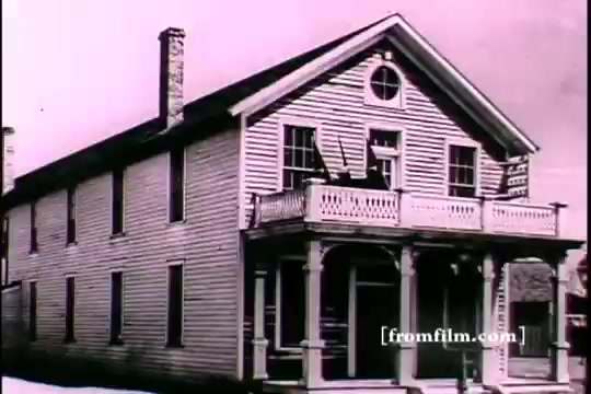 The still shows a historical wooden building, likely a house or small structure, with a distinct architectural style including a peaked roof and a wrap-around porch. The image appears to be in black and white, typical of 16mm prints, conveying a sense of nostalgia. The lack of modern elements enhances its historical context, which relates to Thomas Edison and possibly his connections to early 20th-century American society, including themes of innovation and recruitment.