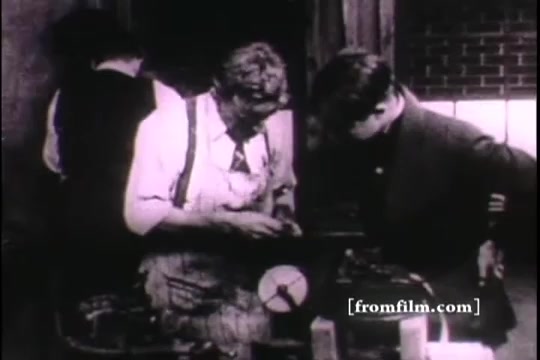 The still from 'Great Moments in History - Thomas Edison - Army Recruiting' depicts a historical scene featuring two individuals engaged in discussion over a piece of machinery, likely related to Edison's work. One person, dressed in work attire, appears to be instructing or demonstrating something to a younger individual. The setting includes industrial elements, indicative of Edison's era, with a backdrop likely representing a workshop or laboratory. The image captures a moment of learning and innovation, reflecting Edison's influence on technology and education.