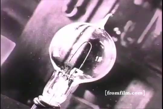 The still features a close-up of an incandescent light bulb, showcasing its clear glass envelope and glowing filament. The image reflects a vintage aesthetic typical of early 20th-century film, embodying the spirit of innovation associated with Thomas Edison. The background is slightly blurred, suggesting a workshop or laboratory setting, emphasizing the theme of invention and progress.