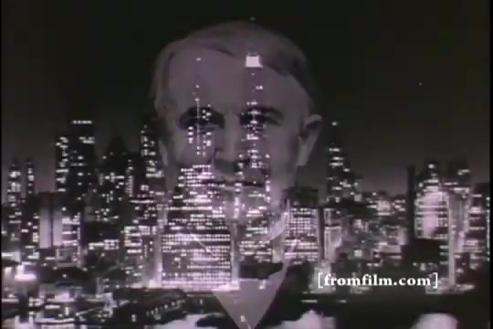 The still features a ghostly double exposure of Thomas Edison’s face merged with a city skyline at night. The urban landscape is illuminated, creating a dramatic backdrop that highlights Edison’s significance. The image evokes a sense of innovation and progress, reflecting the era's fascination with technology and industrial advancement.