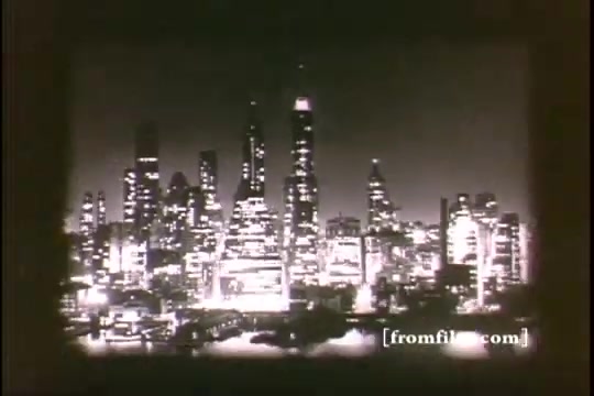 The still from 'Great Moments in History - Thomas Edison - Army Recruiting' features a black-and-white nighttime cityscape, showcasing a skyline filled with towering buildings illuminated by lights. The scene conveys a sense of urban grandeur and historical significance, reflecting an era of innovation associated with Thomas Edison. The focus is on the bustling city atmosphere, indicative of the time period. The image appears to be from a 16mm film print, capturing a nostalgic view of the past.