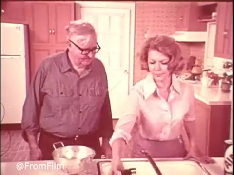The still from the 1970s Ajax Cleanser commercial features a kitchen scene. A man and a woman are engaged in cleaning; the woman is using the product on a sink, showcasing its effectiveness. The tagline 'Turns blue... powers out stains!' suggests the product's stain-fighting abilities. The warm, vintage color palette reflects the era’s style. Follow @FromFilm on Twitter for more commercial clips!