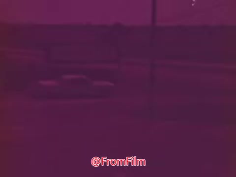 The still from the 'Drive-In' trailer features a blurred scene with a primarily purple tint. It depicts a road with two cars visible in the foreground, likely indicating a drive-in setting. The background appears simplistic and somewhat indistinguishable, emphasizing the focus on the cars. Overall, the still conveys a nostalgic vibe reminiscent of mid-1970s cinema.