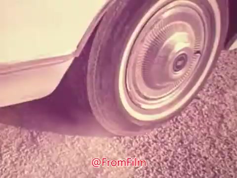 The still from the 1976 trailer for 'Drive-In' focuses on a close-up of a car wheel on gravel. The shot highlights the vintage design of the wheel and tire, conveying a sense of nostalgia typical of the era. The muted colors suggest a stylized or retro aesthetic, indicative of the film's context in the 1970s drive-in culture.