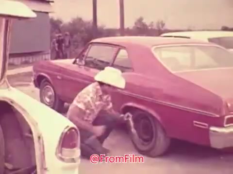 The still from 'Drive-In' (1976) features a scene set in a parking lot, where a man wearing a cowboy hat is crouched beside a red car, appearing to fix a tire. The background shows other vehicles and hints of a casual outdoor environment, capturing a laid-back, nostalgic vibe typical of the era. The colors are soft, contributing to a vintage aesthetic.