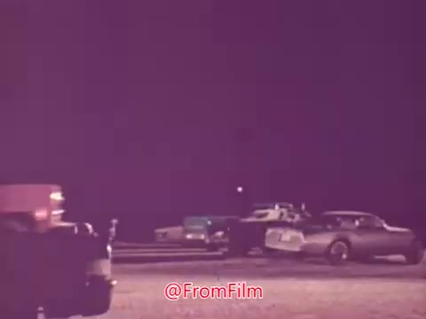 The still from the 1976 movie trailer 'Drive-In' shows a nighttime scene at a drive-in theater. Several vintage cars are parked, with a focus on a line of vehicles facing a screen. The lighting is dim, creating a nostalgic atmosphere typical of that era, highlighting the popularity of drive-ins during the mid-20th century. The film captures a moment in a communal space, likely evoking themes of youth and cinematic escapism.