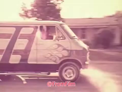 The still depicts a vintage van from the 1976 movie trailer 'Drive-In.' The vehicle features a stylized design with flame motifs and is captured mid-motion, suggesting a dynamic scene. The background is a suburban setting, hinting at a nostalgic vibe typical of the era. The overall aesthetic conveys a playful and adventurous spirit, characteristic of 1970s cinema.