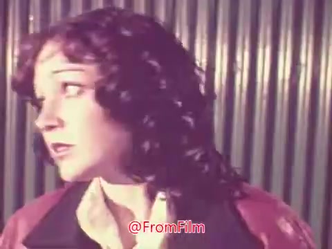 The still from the 1976 trailer for 'Drive-In' features a young woman with curly hair, looking apprehensive or concerned. She wears a colorful jacket, and the background appears to have a corrugated texture, possibly hinting at a drive-in setting. The overall mood suggests tension or suspense, fitting for a trailer context.