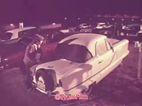 The still likely shows a drive-in movie scene from 1976, featuring a person interacting with a vintage car amidst a parking lot filled with classic automobiles. The color palette suggests a nostalgic vibe, characteristic of the era, emphasizing the drive-in culture and excitement surrounding outdoor movie experiences. The overall atmosphere feels relaxed and captures a moment of connection to the past.