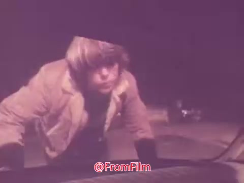 The still features a young man with a distinctive hairstyle, leaning over a car hood in a nighttime setting. Wearing a light-colored jacket, he appears focused, possibly inspecting the vehicle. The ambiance suggests a drive-in theme, fitting with the trailer's context from the 1976 film 'Drive-In.' The color palette has a vintage look, typical of 16mm film.