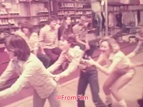 The still from the 1976 movie trailer 'Drive-In' features a group of teenagers engaged in a playful activity, likely a dance or game. They are positioned in a line, with a mix of expressions ranging from joy to concentration. The setting appears to be a fun, lively environment, possibly a roller rink or retro social gathering place, emphasizing the carefree culture of the era. The color tone suggests a vintage aesthetic typical of that time.