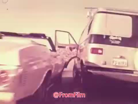 The still from the 1976 movie trailer for 'Drive-In' depicts a dynamic scene featuring two vehicles in a seemingly chaotic moment. A convertible car is closely positioned next to a van with its door flung open, suggesting a sense of urgency or action. The color palette is warm, typical of the 1970s, with a nostalgic aesthetic. The overall composition conveys excitement, likely reflecting themes of adventure or rebellion characteristic of the era's films.