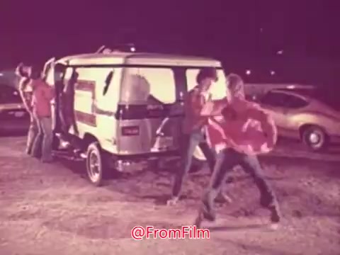 The still from the 1976 movie trailer 'Drive-In' depicts a group of young people interacting near a distinctive van in an outdoor setting. The scene suggests a lively atmosphere, possibly associated with a drive-in movie experience, with vintage cars in the background. The colors appear slightly faded, typical of 16mm film, adding a nostalgic feel to the image. The individuals seem to be engaged in a fun or energetic activity, fitting the youthful theme of the movie.