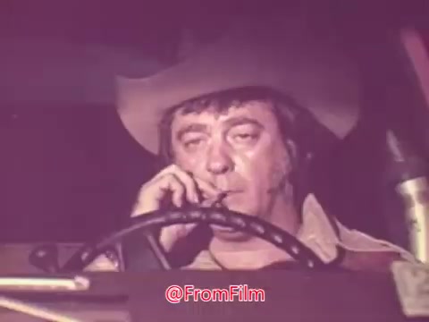 The still from the 1976 trailer for 'Drive-In' features a man sitting in a vehicle, smoking a cigarette. He has a rugged appearance, wearing a cowboy hat, and his expression conveys a sense of contemplation or intensity. The lighting and color tones suggest a vintage aesthetic typical of the era. The steering wheel is prominently visible, creating a confined, introspective atmosphere.