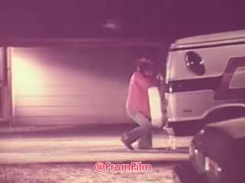 The still captures a nighttime scene from the 1976 film 'Drive-In.' It features a person in a pink shirt and denim pants struggling to hoist a large object next to a parked van, with a garage in the background. The atmosphere suggests a playful or humorous moment, characteristic of the film's lighthearted tone.