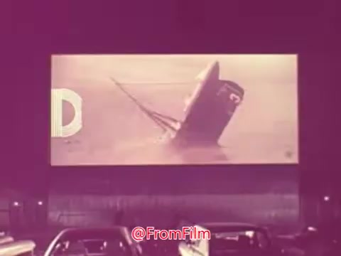 The still from the 1976 movie trailer for 'Drive-In' features a drive-in theater setting, with cars parked in the foreground. The screen displays a dramatic scene of a ship capsizing, suggesting tension and action. The composition captures the nostalgic vibe of the era, with a vintage aesthetic and muted colors.