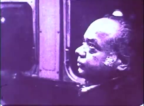 The still depicts a contemplative scene from 'The Laughing Policeman,' featuring a man sitting by a window, possibly in a vehicle or public transport. The lighting creates a somber atmosphere, with a purplish tint typical of older 16mm prints. His expression hints at thoughtfulness or concern, contributing to the film's suspenseful tone. The blurred background suggests movement, enhancing the sense of isolation.