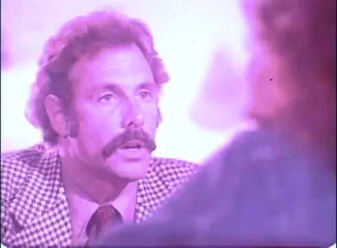 The still from the trailer of 'The Laughing Policeman' features a man with a prominent mustache, dressed in a checkered suit. He appears to be engaged in an intense conversation, with a soft, diffused light casting a pinkish hue over the scene. The focus is on his facial expression, suggesting a moment of high tension or significance in the narrative. The background is slightly blurred, indicating a dialogue-driven moment.