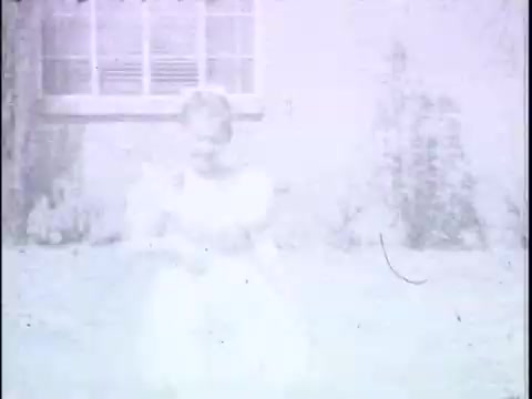 The still appears to show a child in a light-colored dress, possibly interacting with a cat, in a backyard setting. The image is faded and lacks clarity, suggesting age and potential wear from its 16mm film format. It evokes a nostalgic, playful atmosphere typical of early childhood scenes in films.