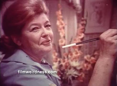 The still features a woman with reddish-brown hair, smiling slightly as she holds a paintbrush. Dressed in a blue outfit, she seems to be engaged in a creative activity. The background includes floral decorations, contributing to a warm, cozy atmosphere typical of 1970s aesthetics. The image conveys a sense of comfort and inviting creativity, in line with the theme of the Ben Gay commercial portraying warmth and relief.