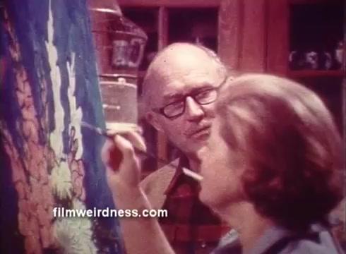 The still from the 1970s Ben Gay commercial depicts two individuals engaged in a creative process. The male figure, wearing glasses and a checkered shirt, observes attentively as a female figure paints on a colorful canvas. The atmosphere suggests a warm, collaborative environment, reflecting the soothing qualities associated with the Ben Gay product. The setting appears homely, with wooden cabinetry in the background, typical of the era's aesthetic.
