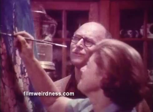 The still depicts a scene from the 1970s Ben Gay commercial. It features two individuals, likely in a cozy, homey environment, engaged in a creative activity, possibly painting. The focus seems to be on collaboration and warmth, aligning with the advertisement's theme of comfort and relief offered by Ben Gay. The setting includes elements typical of the era, enhancing the nostalgic vibe.