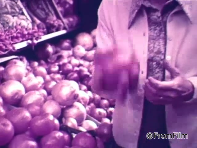 The still from the 1960s Chef Boyardee spaghetti sauce commercial shows a person in a grocery store, surrounded by an abundance of fresh produce, particularly onions. The individual appears to be emphasizing the quality of the ingredients essential for making good spaghetti sauce, suggesting a tactile evaluation by handling the vegetables. The vibrant, slightly vintage color palette contributes to the nostalgia associated with this era of food advertising, highlighting the importance of fresh, quality ingredients.