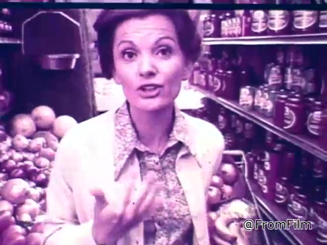 The 16mm still from the 1960s Chef Boyardee spaghetti sauce commercial features a woman in a grocery store. She engages the audience with an enthusiastic expression, discussing how to select quality spaghetti sauce. In the background, jars of Chef Boyardee sauce are visible alongside fresh produce, including apples. The scene conveys a focus on fresh ingredients and highlights the brand's product as a reliable choice for consumers.