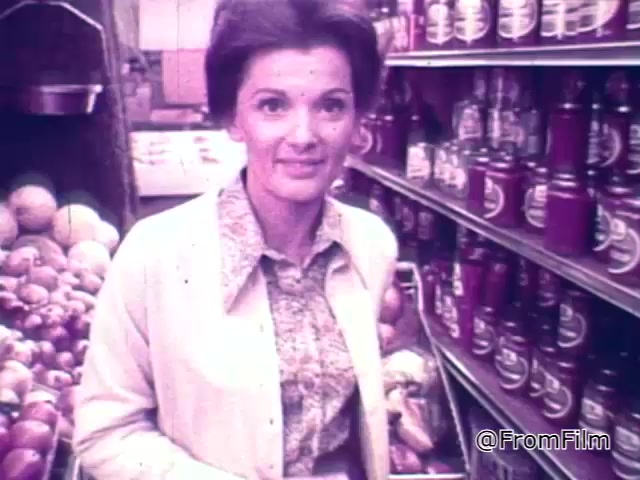 The 16mm still from the 1960s Chef Boyardee spaghetti sauce commercial features a woman smiling in a grocery store aisle. She is dressed in a light-colored jacket with a patterned blouse underneath. Behind her, shelves are lined with jars of Chef Boyardee spaghetti sauce, alongside a display of fresh produce like onions and melons. The overall aesthetic is warm and nostalgic, capturing the shopping experience of that era. The voiceover emphasizes the importance of quality ingredients in cooking, transitioning into the merits of selecting Chef Boyardee for spaghetti sauce.