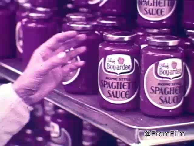 The still depicts a nostalgic grocery store scene from a 1960s Chef Boyardee spaghetti sauce commercial. A hand reaches toward jars of Chef Boyardee spaghetti sauce, prominently displayed on a shelf. The jars feature a classic design, with the brand name and product type clearly visible on the labels. The warm colors and retro aesthetic evoke a sense of the era, highlighting the appeal of convenience in home cooking. The voiceover emphasizes the quality of ingredients in spaghetti sauce, suggesting that viewers choose based on visual richness and texture.