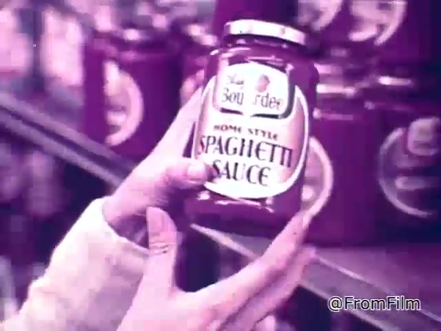 In this still from a 1960s Chef Boyardee spaghetti sauce commercial, a hand is prominently holding a jar of Chef Boyardee Home Style Spaghetti Sauce. The jar features a classic design, with the brand name clearly visible. In the background, several similar jars are displayed on store shelves, emphasizing the product's availability. The overall tone of the image is nostalgic, reflecting the advertising style of that era, focused on the quality and visual appeal of the sauce.