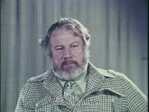 The still features Peter Ustinov, a distinguished actor and humanitarian, delivering a message in a UNICEF public service announcement from 1976. He appears contemplative, with a well-groomed beard, wearing a patterned jacket and tie. The backdrop is neutral, focusing attention on his expression and the seriousness of the topic he addresses, likely related to children's welfare and humanitarian issues.