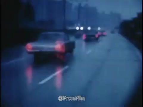 The still from the 1968 National Safety Council PSA conveys a moody, atmospheric scene. It captures a rainy road where several cars are navigating, with soft reflections of their headlights illuminating the wet pavement. The colors appear muted, emphasizing the somber effect of the rain and the potential hazards of driving under such conditions.   The message highlights the dangers of driving with worn tires, excessive speed, and in adverse weather, portraying the road as a concealed trap waiting for the unwary driver. The imagery reinforces the PSA's message about the importance of being cautious and maintaining vehicle safety to avoid accidents.