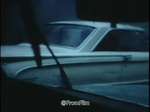 The still from the 1968 National Safety Council PSA captures a scene that conveys a sense of caution and alertness regarding driving conditions. The image features a close-up view from inside a car, where rain is visibly falling, creating a blurred effect on the windshield. Outside, another vehicle, likely moving slowly due to the slippery conditions, is partially shown, emphasizing the theme of road safety amid adverse weather.  The color palette is subdued, indicative of a rainy day, which adds to the somber tone of the message about the potential dangers of driving in such conditions. The presence of rain enhances the idea of hazards like skids and loss of control. The still effectively embodies the PSA's warning about the importance of maintaining proper tire wear and pressure, alongside