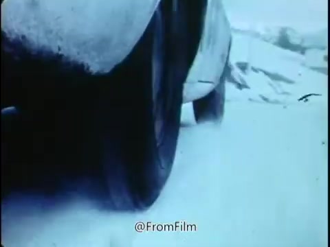 The still from the 1968 National Safety Council PSA depicts a close-up of a tire on a vehicle navigating through snow. The tire appears to be in motion, emphasizing the theme of safety and caution in adverse weather conditions. The overall tone suggests a warning about the dangers of inadequate tire maintenance, such as tread wear and incorrect tire pressure, especially in slippery conditions. The visuals convey a sense of urgency and alertness, reinforcing the message to be conscious of potential hazards while driving.