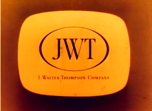The still features the logo of the J. Walter Thompson Company (JWT), a prominent advertising agency, rendered in a retro style. The logo is displayed within an oval frame, with the initials 'JWT' prominently featured. The color scheme is warm, with orange and yellow tones, typical of the era. This frame is part of the silent 16mm Kodak sales film from 1977, likely aimed at promoting Kodak products during the Christmas season, possibly with contributions from notable figures Michael Landon and Dick Van Dyke.