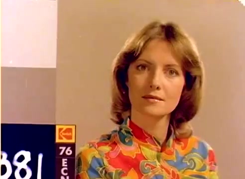 The still features a young woman with medium-length hair, styled in soft waves, dressed in a vibrant, multicolored floral shirt. She gazes intently at the camera, conveying a sense of poise. In the foreground, there is a Kodak color chart, indicating it is from a 16mm silent negative. The context suggests this frame is part of a 1977 sales film associated with the festive season, showcasing Kodak's products and likely featuring Michael Landon and Dick Van Dyke in other segments.