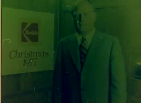 The still depicts a man in a suit standing in front of a Kodak sign that reads 'Christmas 1977.' The image is from a 16mm silent color negative, likely featuring promotional content for Kodak's products during the holiday season. The color has a greenish tint, characteristic of aging film, and there are some visible artifacts typical of older film stock.