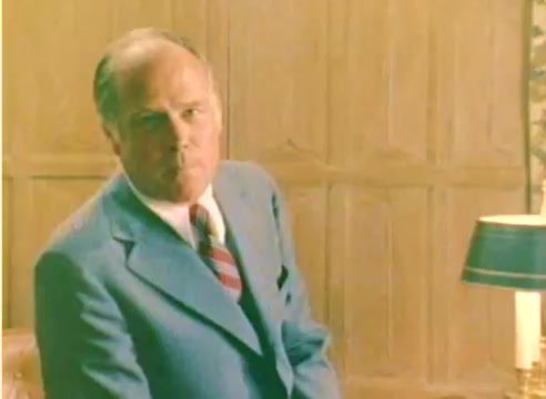 The still features a man in a light blue suit, seated with a serious expression. The background includes wood paneling and a lamp, suggesting an indoor setting. This is a scene from a Kodak 1977 sales film, highlighting the season's promotional efforts, showcasing a professional and polished presentation.