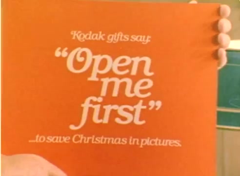 The still from the 'Kodak 1977 Sales Film' features a hand holding an orange card with the text, 'Kodak gifts say 'Open me first' ... to save Christmas in pictures.' The design emphasizes the urgency and importance of Kodak products as essential for capturing holiday memories. The color and playful font reflect the festive spirit of the 1977 Christmas season.