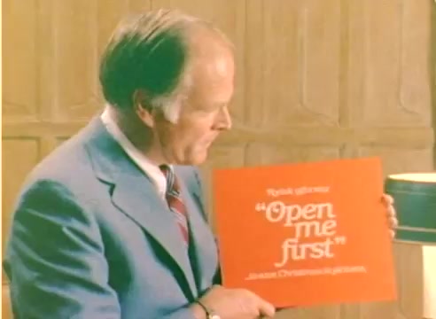The still from the 'Kodak 1977 Sales Film' features a man in a suit, holding a bright orange card that reads 'Open me first.' The background is a simple, wooden setting, creating a warm, nostalgic atmosphere. This scene likely aims to promote Kodak's products in anticipation of the Christmas season. The film is silent and shot in color, capturing a moment of anticipation and festive spirit.