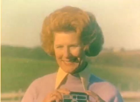 The still features a woman with curly, reddish hair, smiling joyfully while holding a camera. She wears a light purple sweater over a collared shirt. The background shows a blurred landscape, typical of the muted colors found in 16mm film from the 1970s. The overall vibe is nostalgic, reflecting the era’s style and the context of a Kodak sales film aimed at promoting photography during the holiday season.