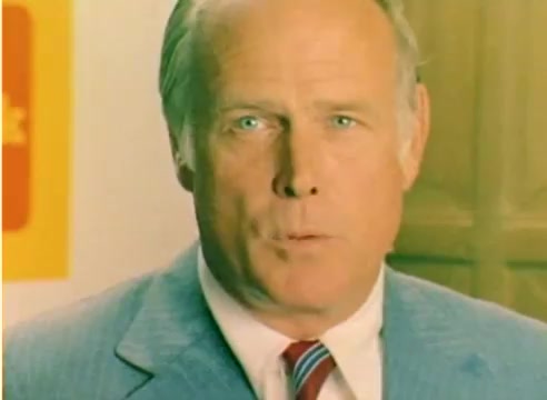 The still from the 'Kodak 1977 Sales Film' features a well-dressed man with light-colored eyes, speaking directly to the camera. The background suggests a promotional setting, typical of marketing visuals from the era. The film likely emphasizes Kodak products in the context of the approaching Christmas season, utilizing a warm color palette typical of color negatives from the 1970s. The overall tone is professional and engaging, aiming to captivate the audience for a holiday sales pitch.