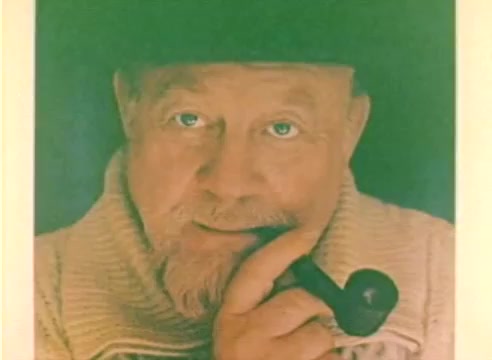The still features a close-up of a man with a beard and a pipe, wearing a light-colored sweater and a hat. The image has a warm color palette that conveys a nostalgic feel. This production, a silent color negative, is associated with the 1977 Christmas season and highlights the promotional efforts of Kodak. The context suggests a blend of charm and warmth typical of holiday-themed media.