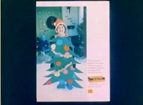 The still from the 'Kodak 1977 Sales Film' features a young child dressed in a festive costume resembling a Christmas tree, complete with colorful decorations. The background shows a decorated room, likely in preparation for the holiday season. The muted colors and overall presentation reflect the nostalgic vibe of the era, emphasizing Kodak's focus on capturing cherished moments during Christmas. The image is part of a silent 16mm negative, showcasing the festive spirit leading up to the holidays.