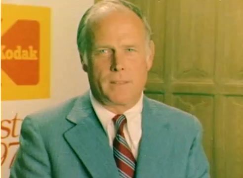 The still from the 'Kodak 1977 Sales Film' features a well-dressed man in a light blue jacket and red-striped tie, standing in front of a Kodak logo. The background includes wooden paneling, suggesting a warm, inviting atmosphere. It showcases promotional content aimed at the Christmas season, typical of Kodak’s marketing approach during that era. The colors and clarity reflect the characteristics of a 16mm silent color negative.
