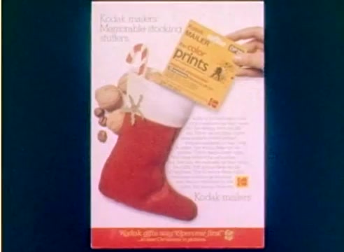 The still features a festive red Christmas stocking overflowing with various holiday treats, including candy canes and nuts. Above the stocking, a hand is displaying a Kodak product box labeled 'Holiday Color Prints.' The background is a simple, light color, enhancing the focus on the stocking and the Kodak advertisement, emphasizing memorable holiday moments. The overall tone is cheerful and promotional, aligning with the lead-up to the 1977 Christmas season.